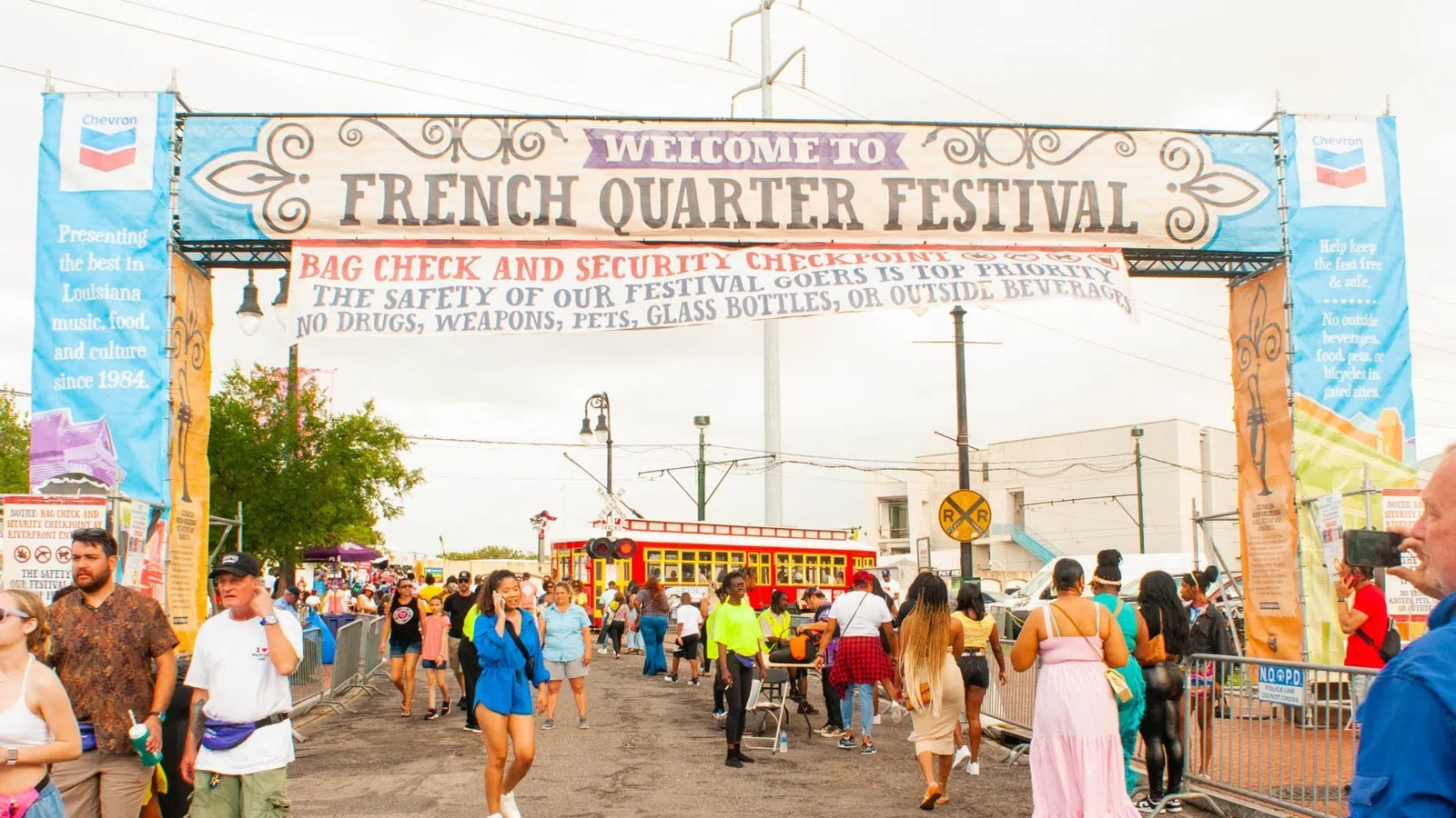 Crowd entering French Quarter Festival under welcome banner with security check and streetcar in background