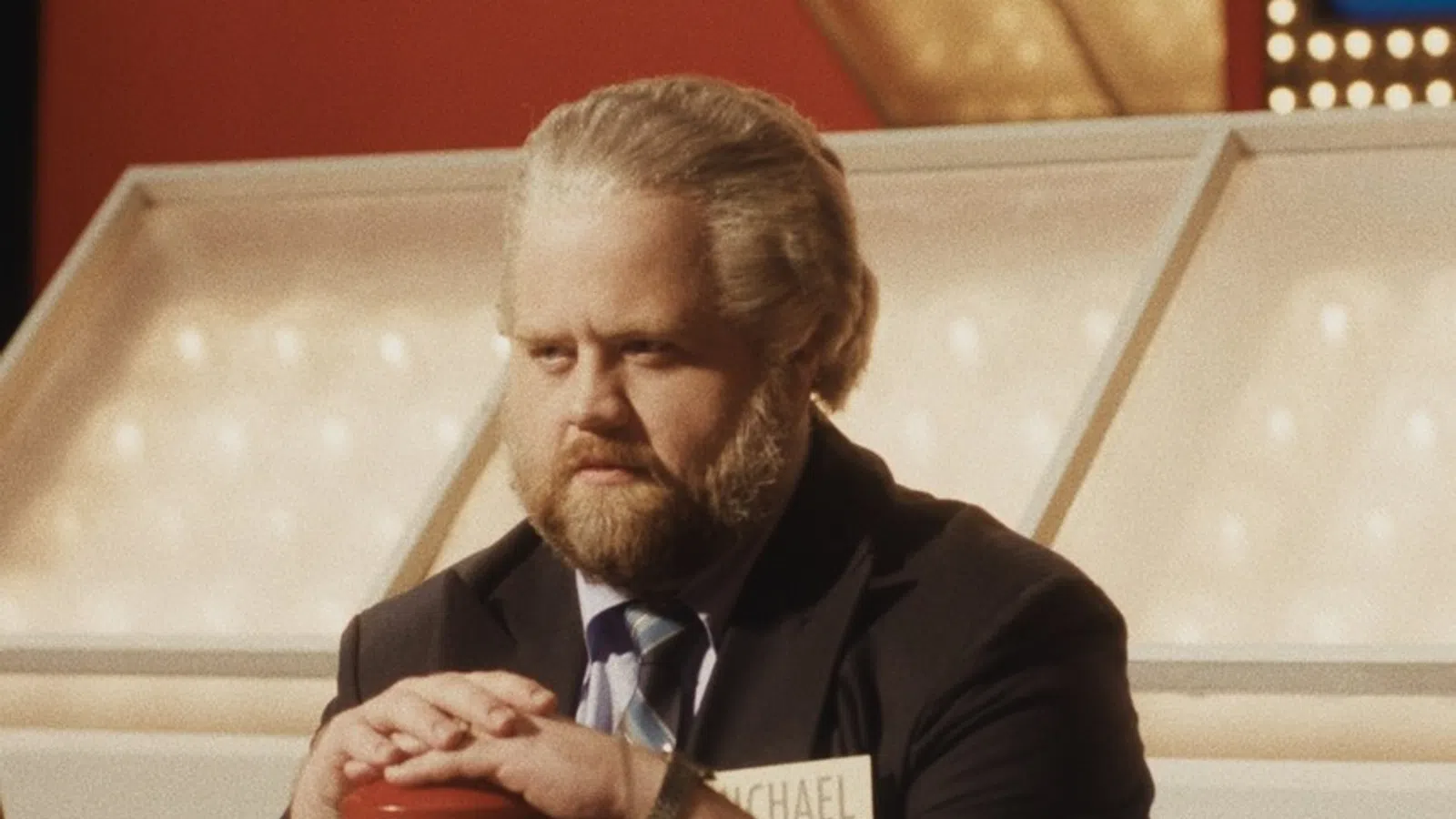 Man with beard and suit pressing a buzzer on a game show set with name tag reading Michael.