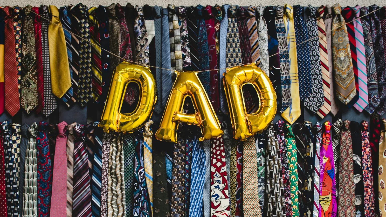 Gold balloons spelling DAD hang among rows of colorful patterned men's neckties on a wall display.