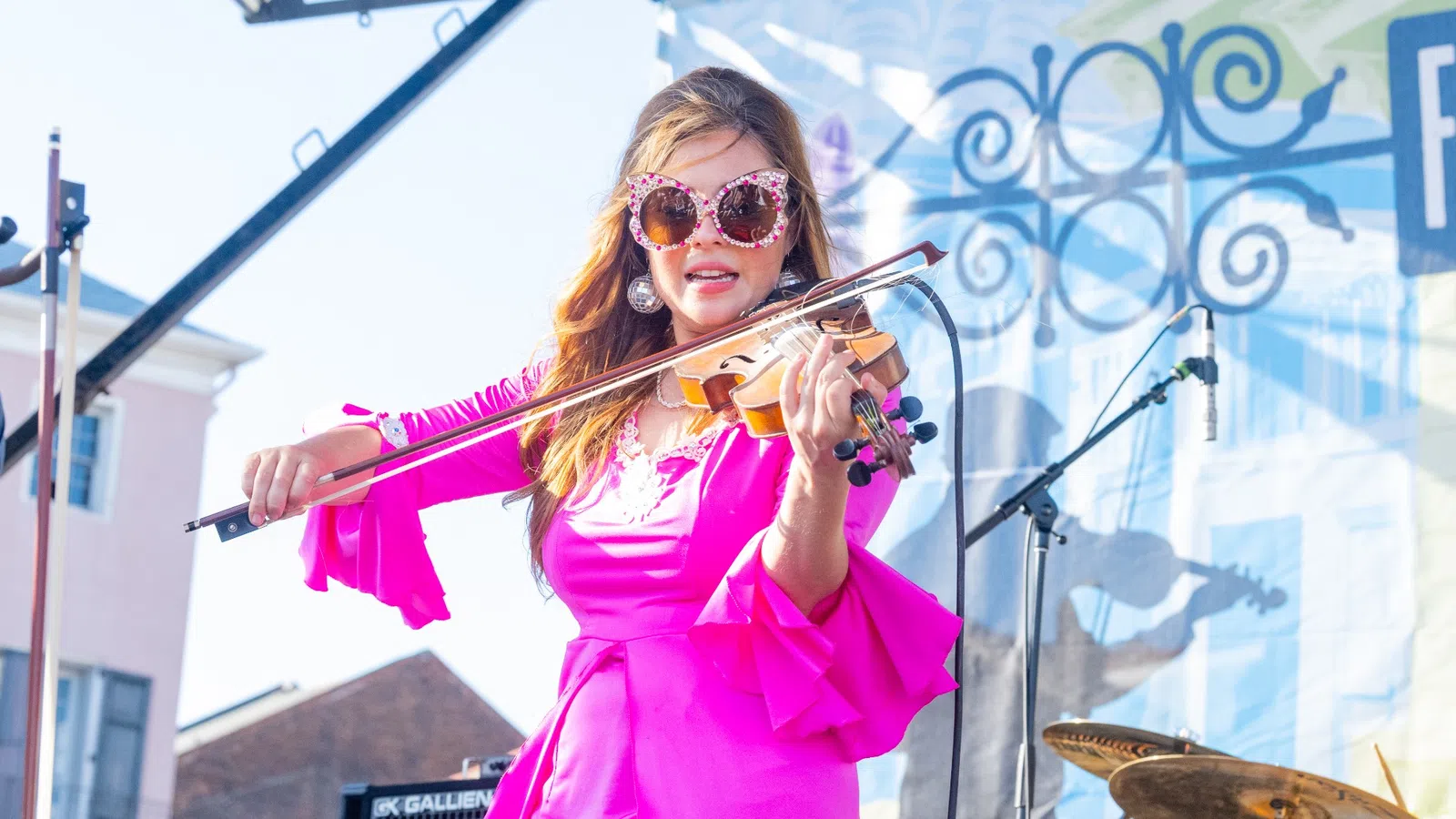 Woman in bright pink dress playing violin on outdoor stage during daytime musical performance.