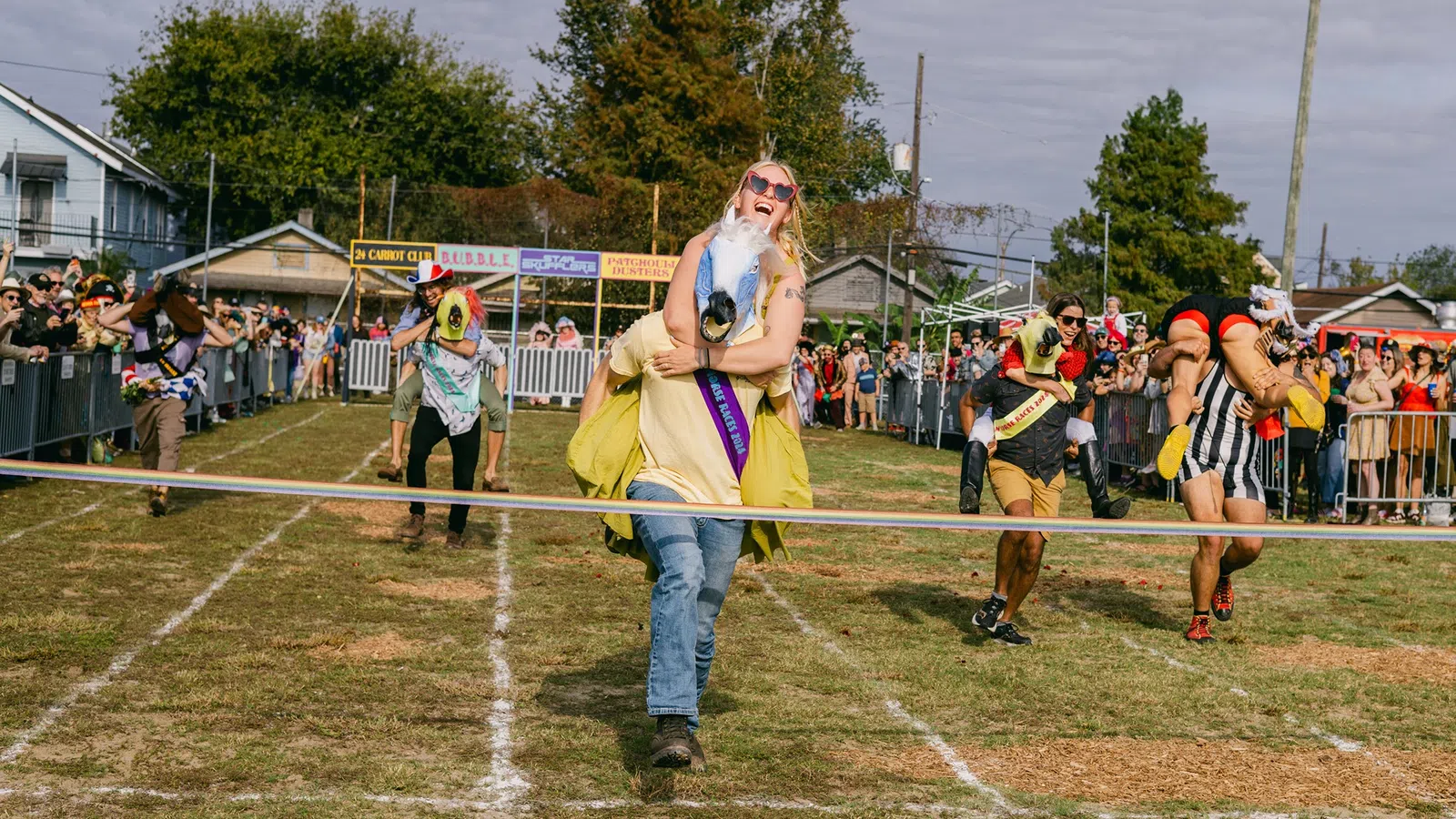 Couples race in a horse costume event on a grass track with spectators cheering behind barriers.