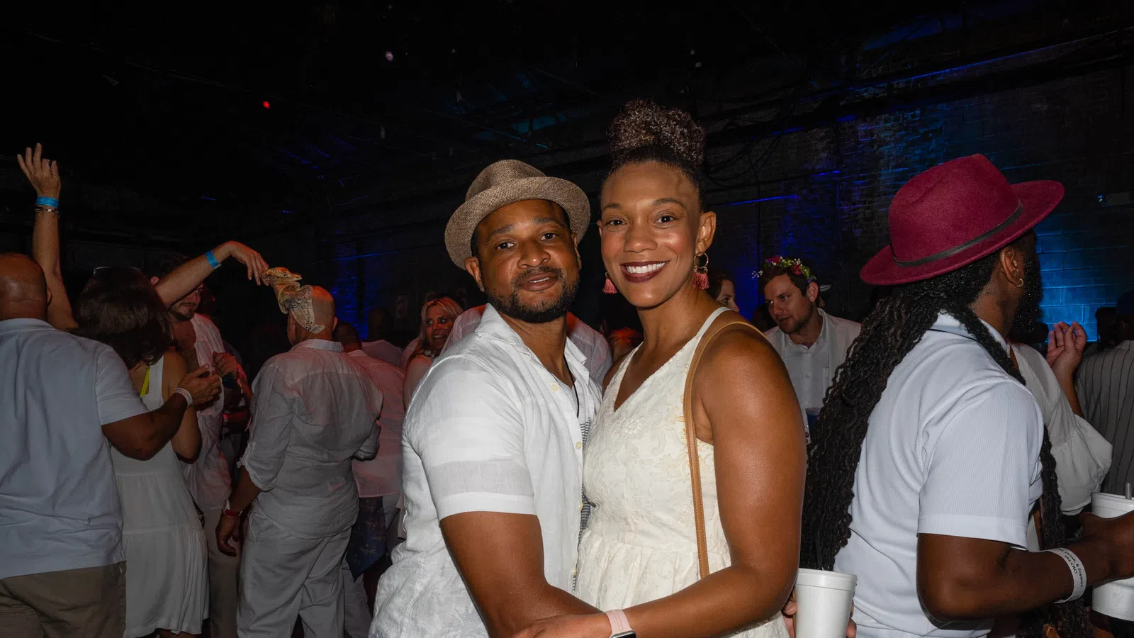Couple in white outfits smiling and embracing at a lively indoor party with colorful lighting and crowd.