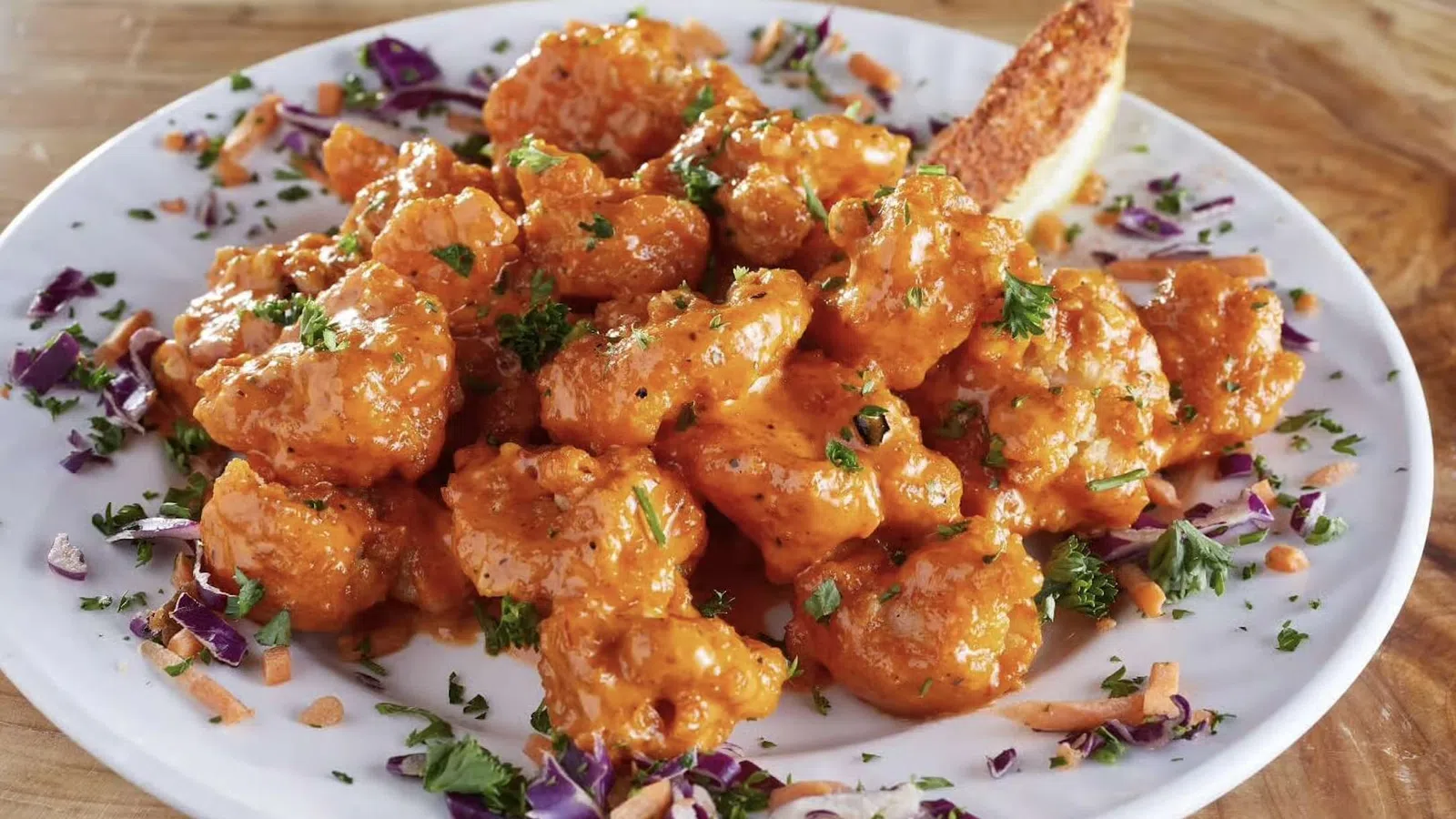 Buffalo cauliflower bites garnished with parsley and shredded vegetables on a white plate with a breadstick.