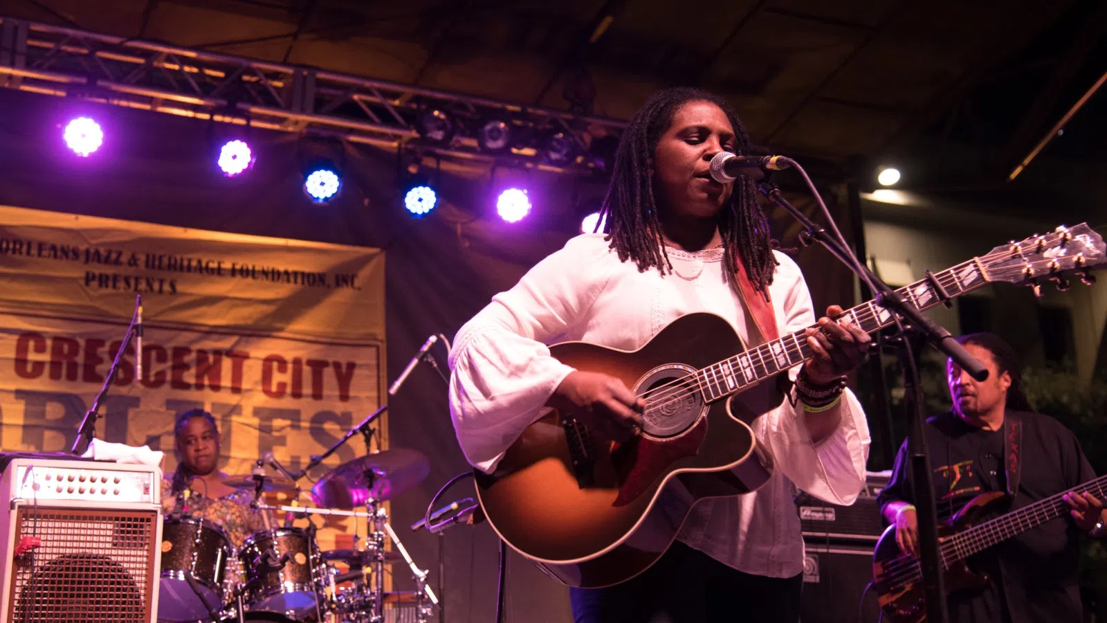 Female musician singing and playing guitar on stage with band at Crescent City Blues festival under purple lights.