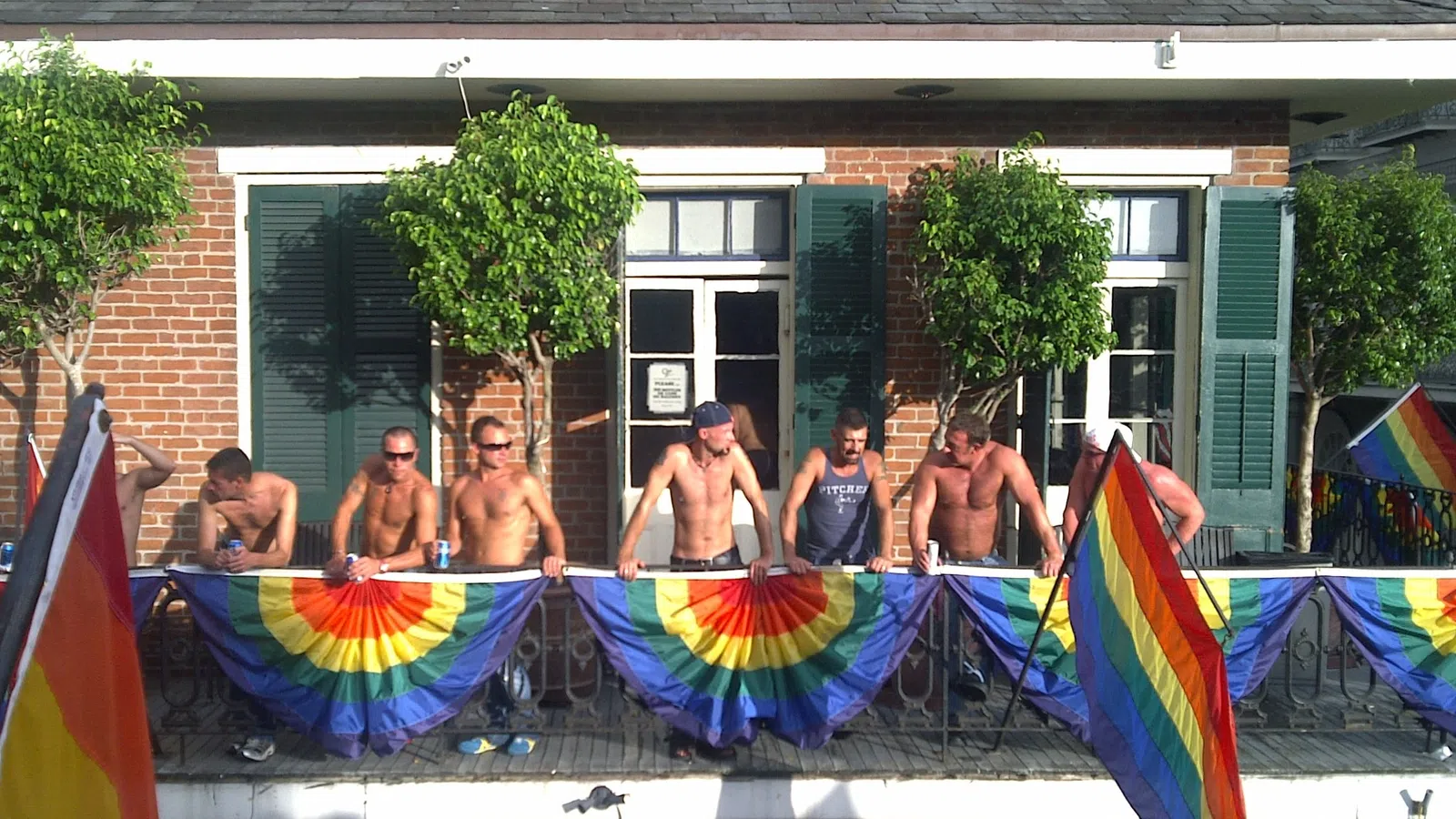 Group of shirtless men holding rainbow flags on a balcony outside a brick building under sunny sky