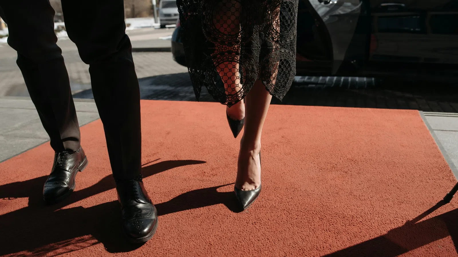 Man and woman walking on a red carpet, showing black shoes and woman in a lace dress.
