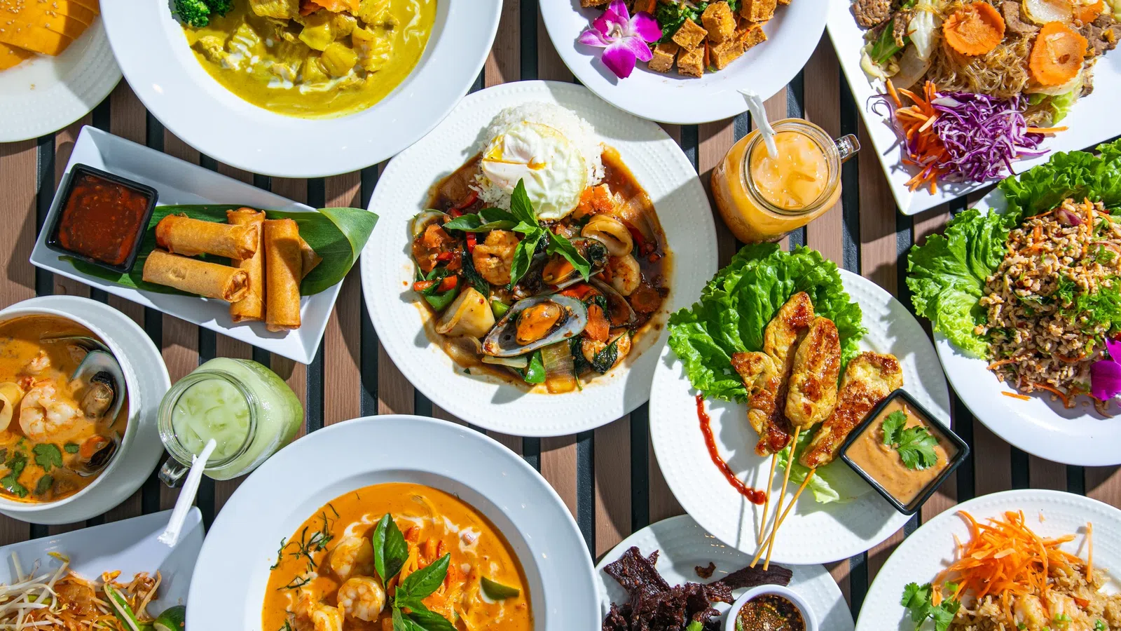 Top-down view of diverse Thai dishes including curry, spring rolls, grilled skewers, seafood, and fresh vegetables on wooden table.