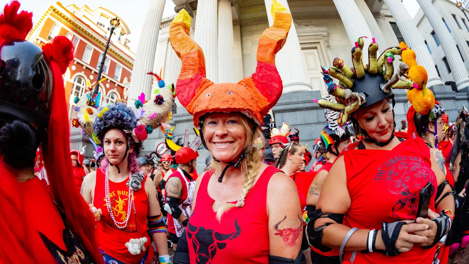 Group of people in red shirts and elaborate horned helmets gathered outside a historic building with columns.