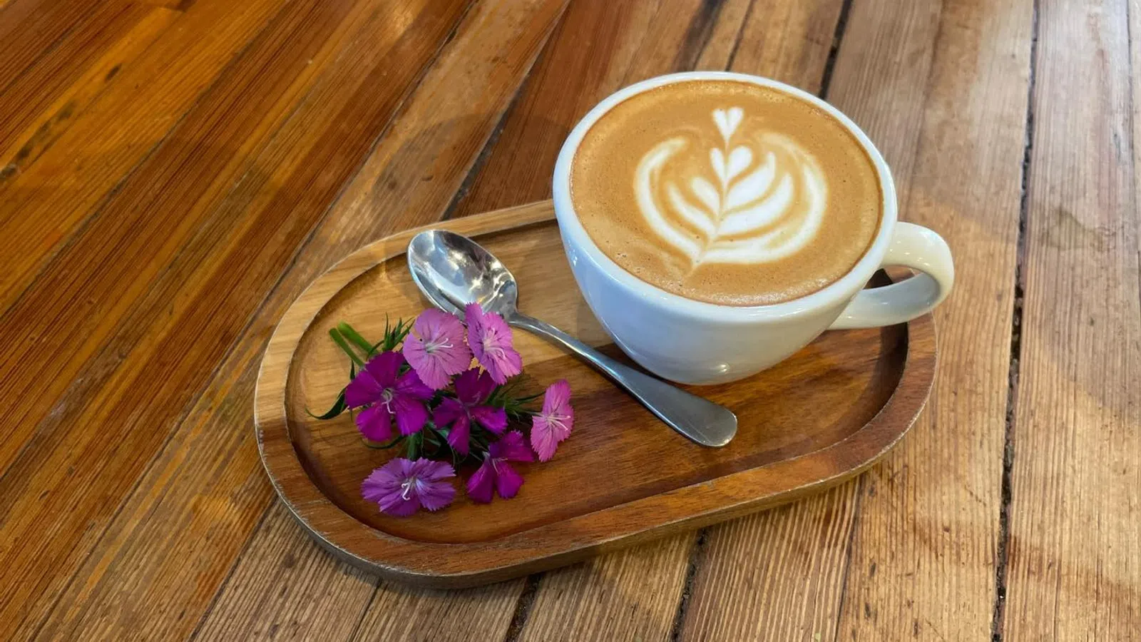 White cup of latte with heart-shaped latte art on a wooden platter with purple flowers and spoon.