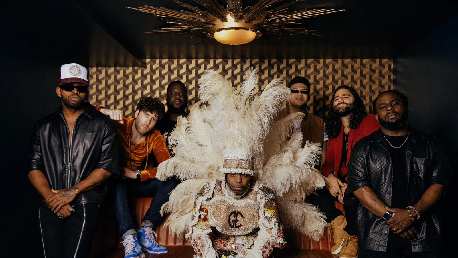 Group of seven men posing indoors with one in an elaborate feathered costume and others in casual and stylish attire