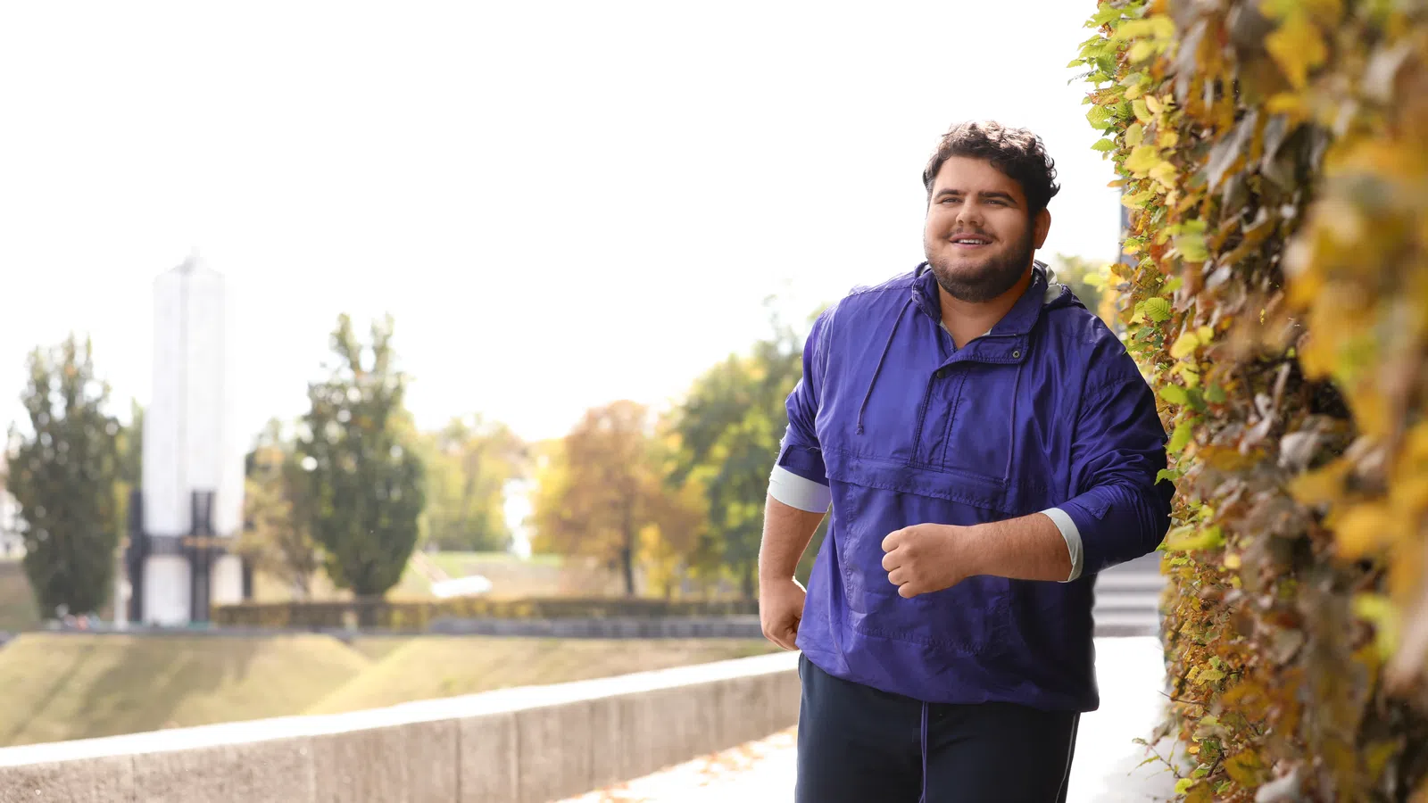 Smiling man in purple jacket jogging outdoors along a wall with autumn foliage in the background.