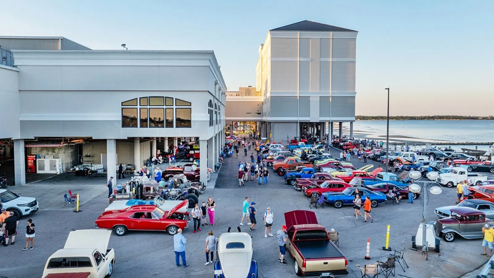 Outdoor classic car show with vintage cars and attendees near waterfront buildings at sunset.
