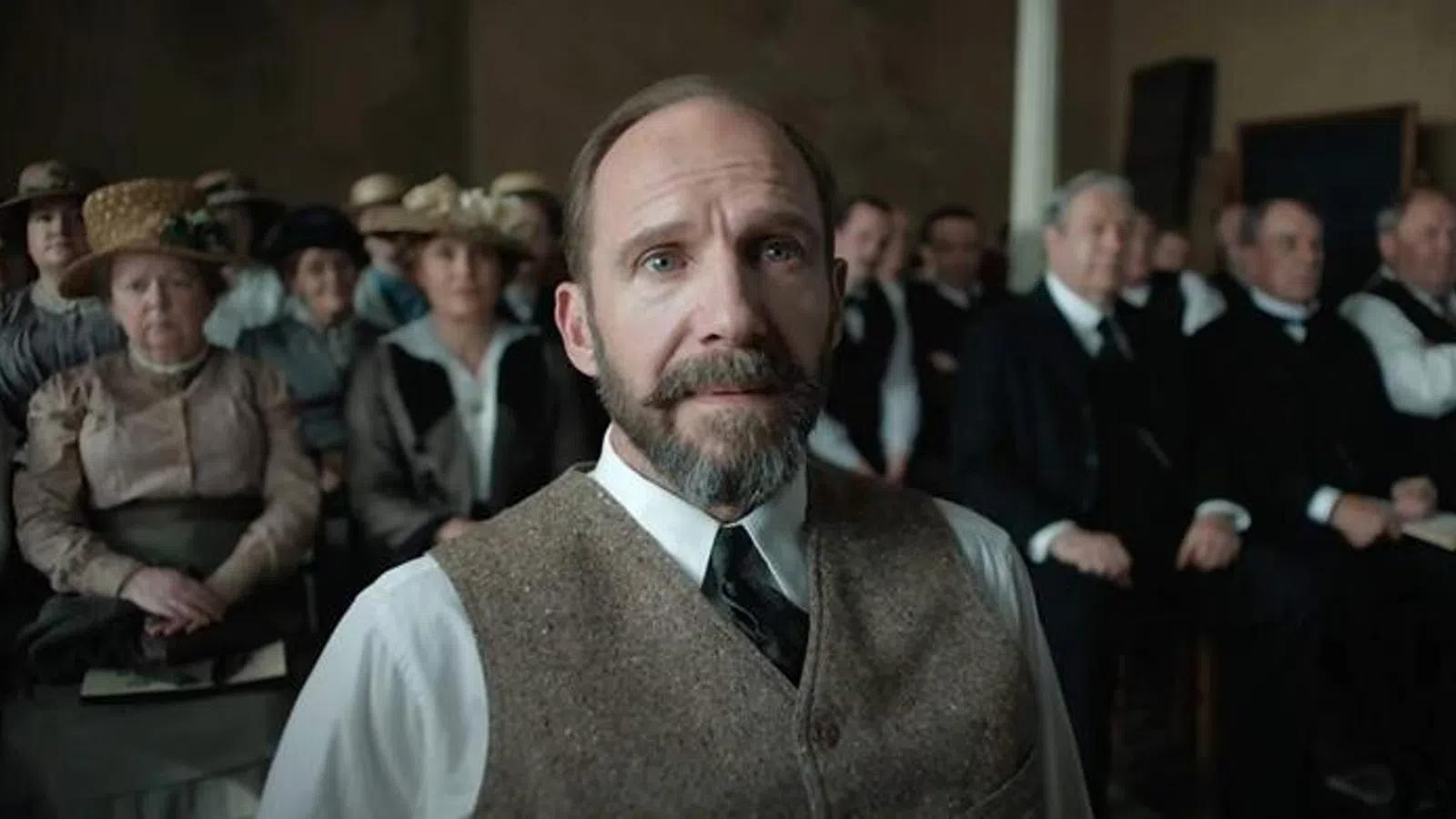 Man with beard and vest sitting attentively in a vintage courtroom with formally dressed audience behind him.