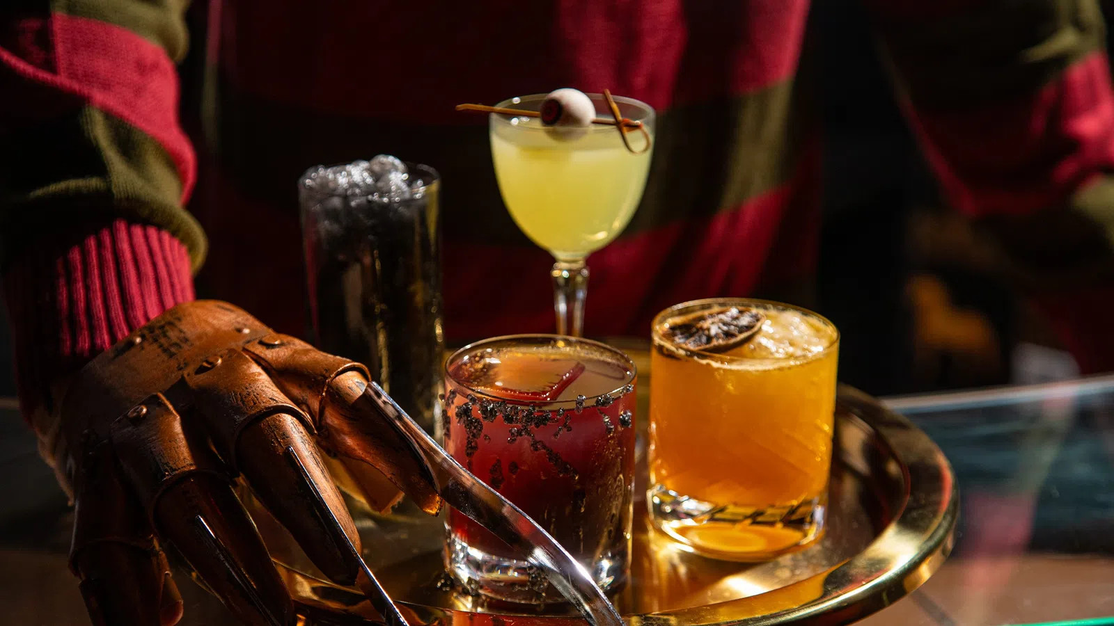 Person in striped sweater and clawed glove serving four colorful cocktails on a round tray at a bar