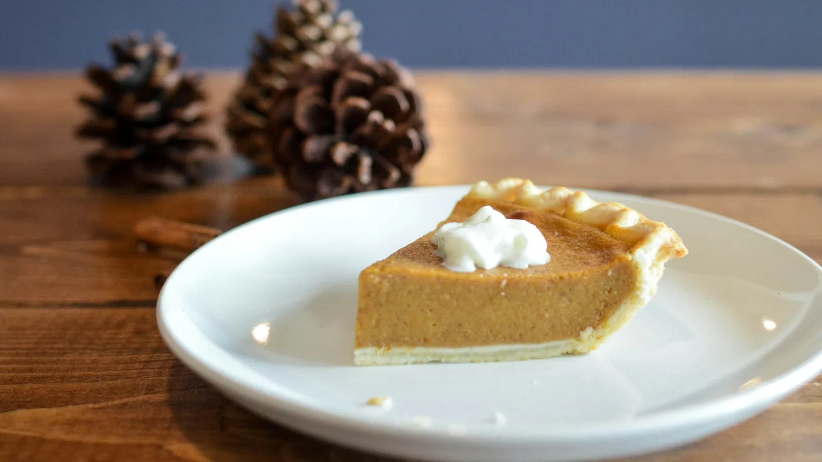 Slice of pumpkin pie with whipped cream on white plate with pine cones on wooden table.