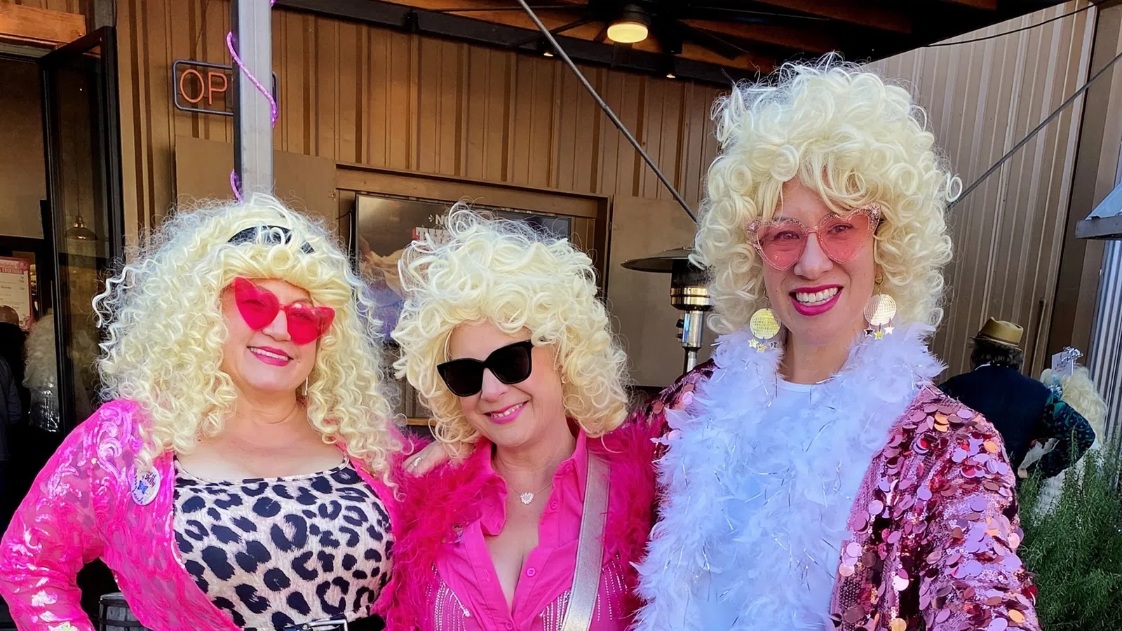 Three women wearing blonde curly wigs and vibrant pink outfits with sunglasses, smiling and posing outdoors.