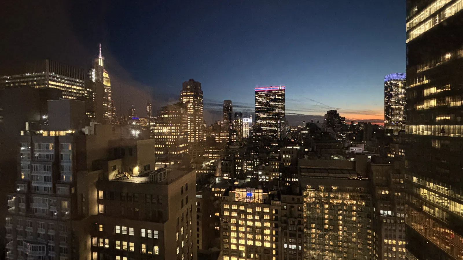 City skyline at dusk with illuminated skyscrapers and a clear fading sunset in the background.