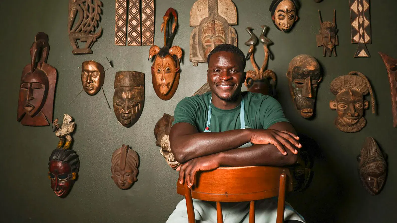 A smiling man poses with African masks hanging on a wall, showcasing cultural art and heritage.