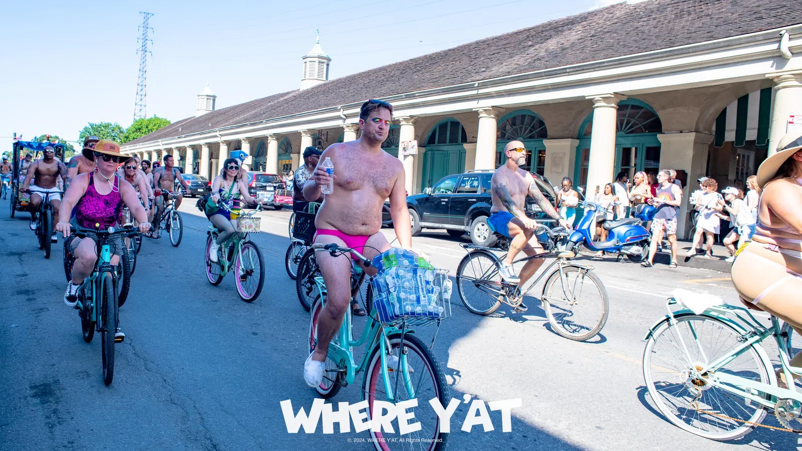 Group of minimally clothed cyclists riding on a sunny street during a festive event with spectators watching