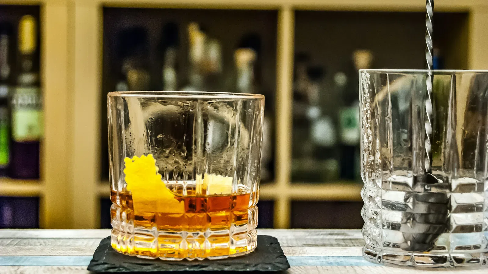 Whiskey glass with orange peel on black coaster next to empty mixing glass with stirring spoon on wooden bar counter.