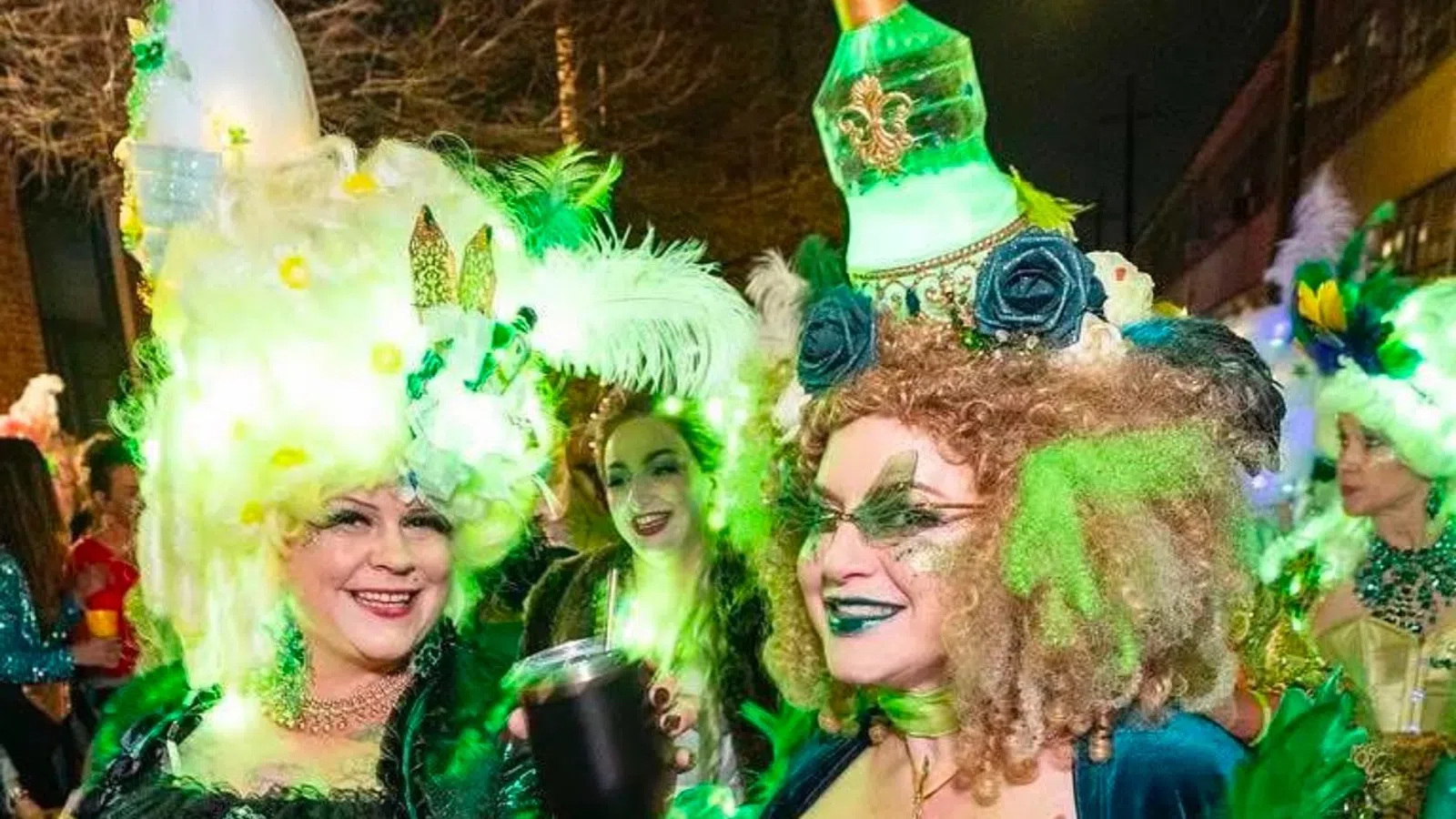 Two women in elaborate, glowing green Mardi Gras costumes with feathers and decorative headpieces at night