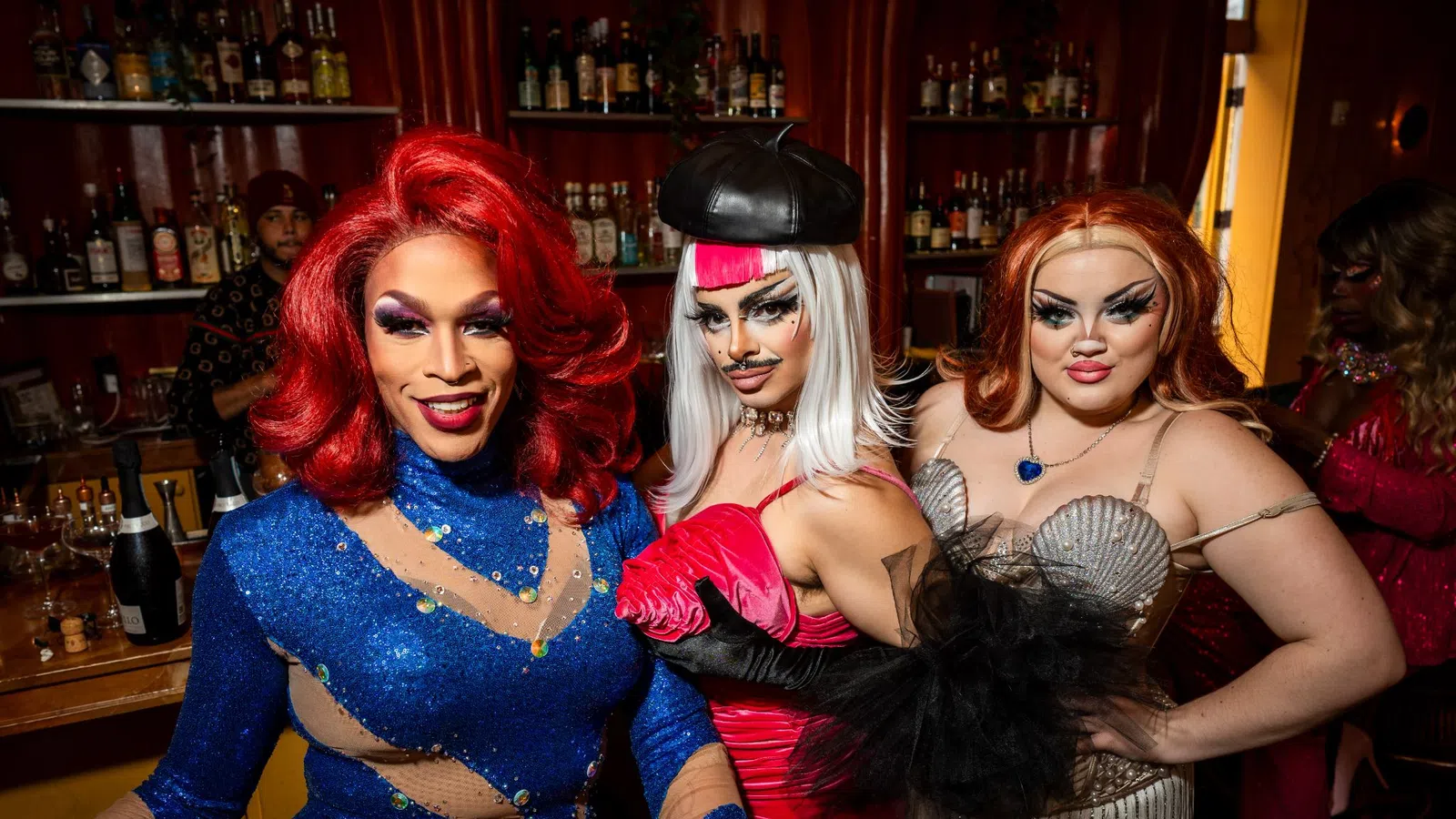 Three drag performers in colorful glamorous outfits pose confidently inside a bar with liquor shelves in the background.