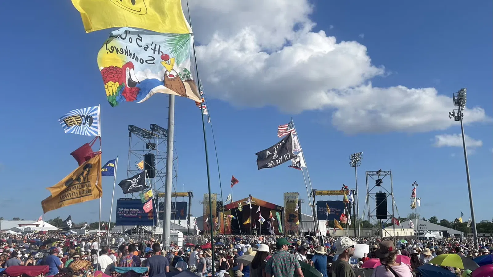 A vibrant festival scene with colorful flags and a large crowd enjoying outdoor music under a blue sky.