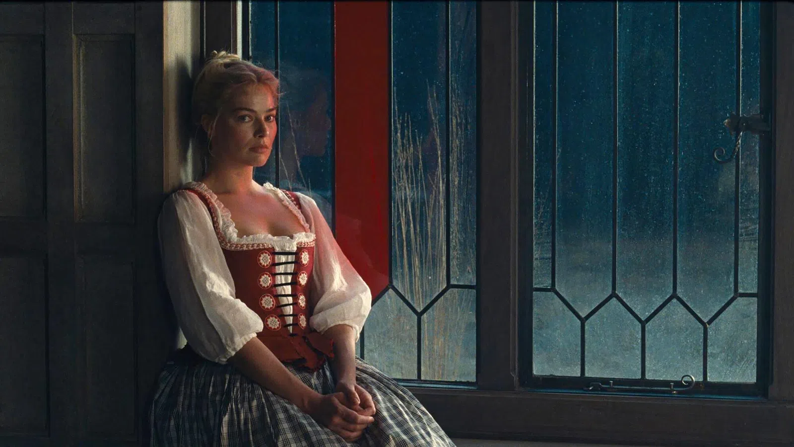 Woman in vintage dress sitting pensively by a large window with stained glass panes in dim light