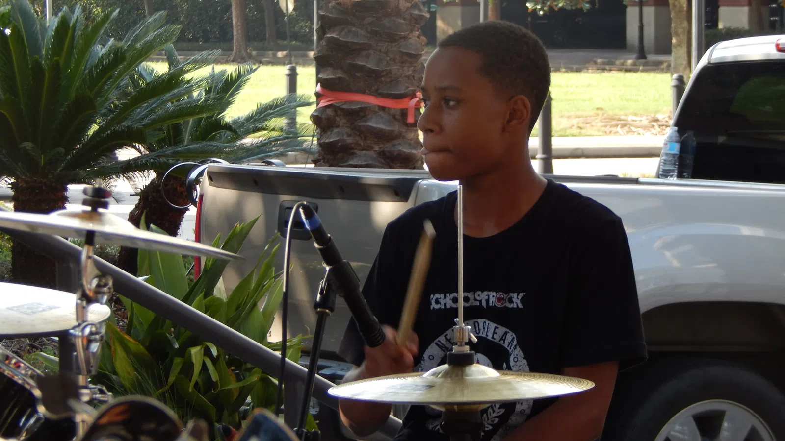 Young boy playing drum set outdoors during the daytime beside greenery and parked cars.