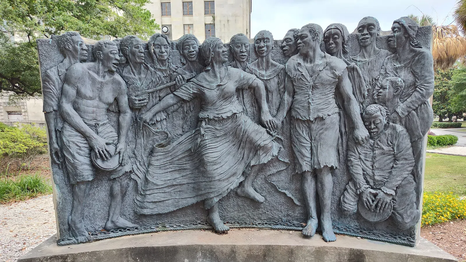 Bronze relief sculpture of joyful people dancing and playing drums in an outdoor park setting.