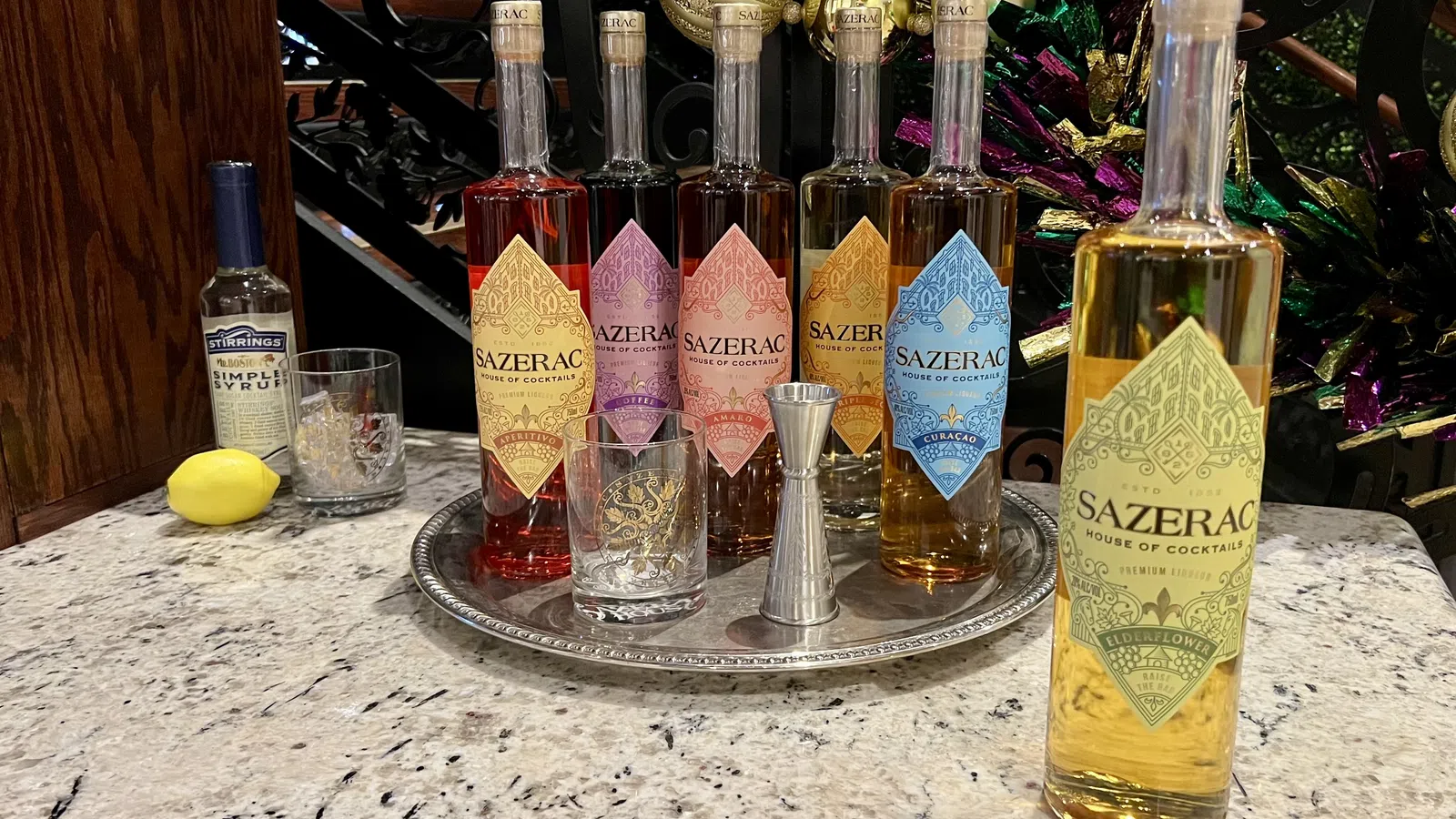 Six colorful Sazerac cocktail liquor bottles arranged on a silver tray with glasses, jigger, syrup, and a lemon on marble countertop.
