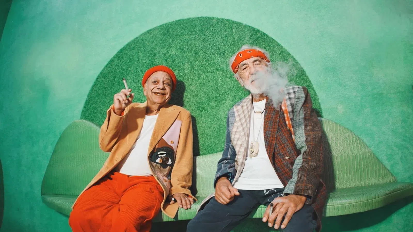 Two stylish seniors in vibrant outfits relaxing on a green bench with smoke in the air.