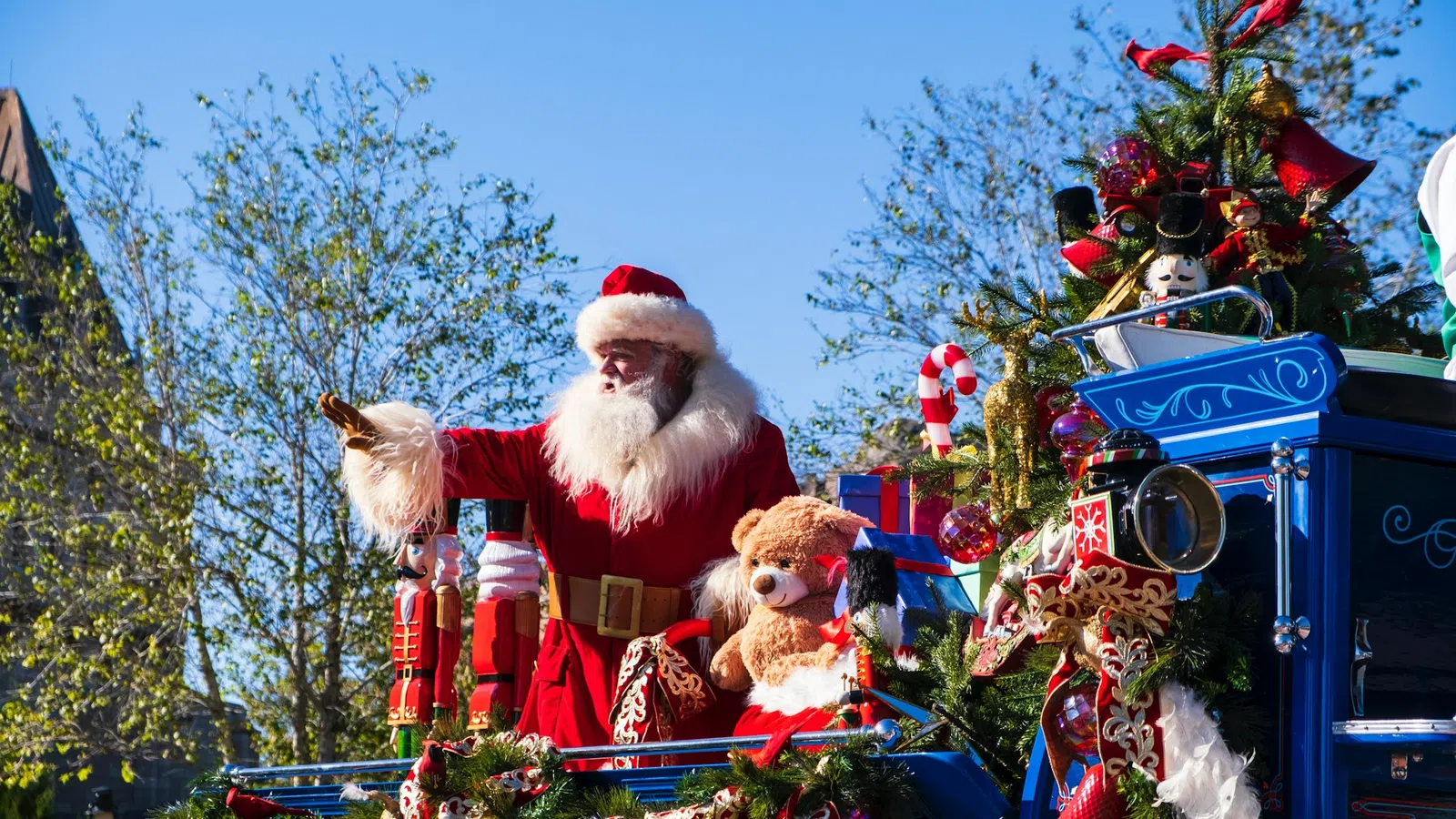 Santa Claus waves from a festive blue parade float decorated with toys, Christmas garlands, and candy canes under clear skies.