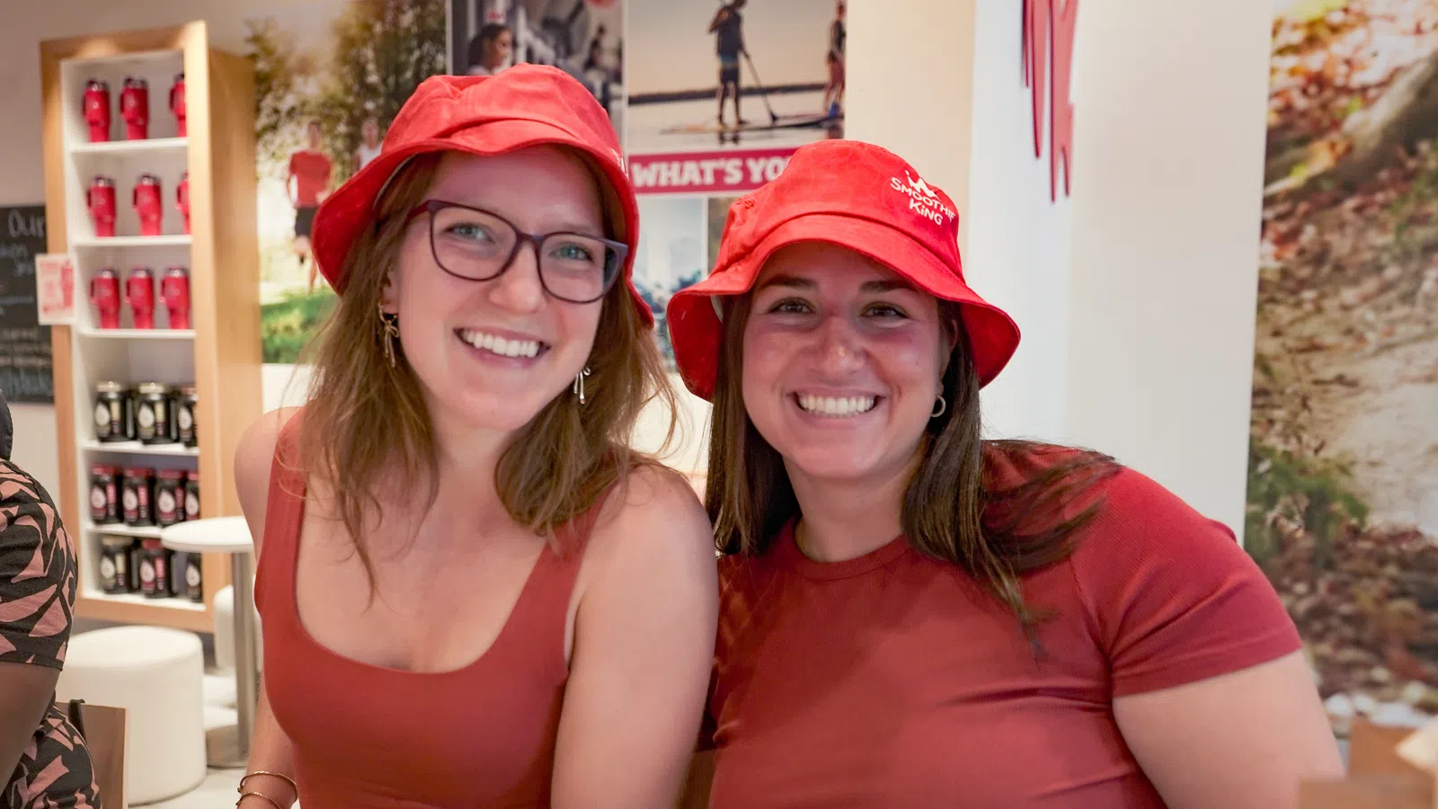 Two smiling women wearing red shirts and red hats sitting indoors with shelves and posters in the background