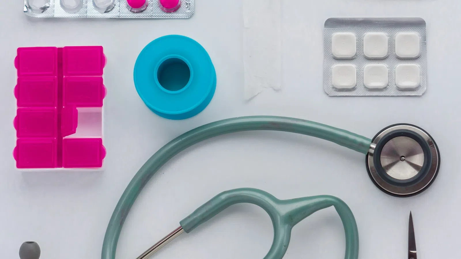 Medical supplies including stethoscope, pill organizer, bandages, tablets, and medical tape on a white surface.