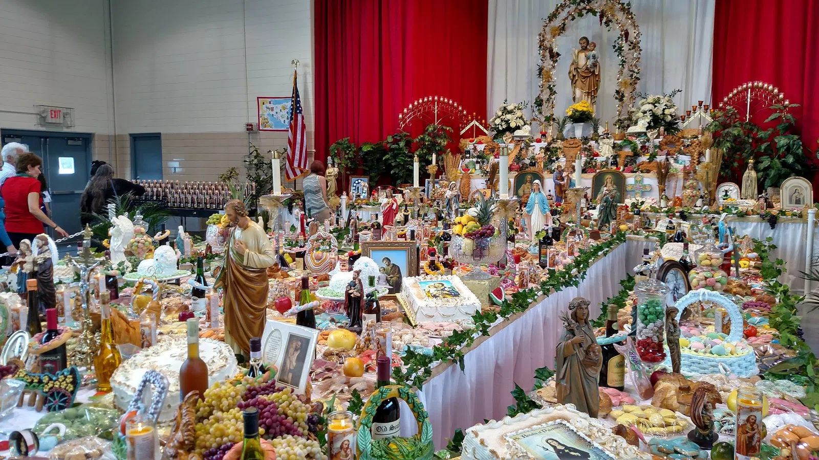 Indoor religious altar with statues, candles, flowers, food offerings, and people attending a celebration.