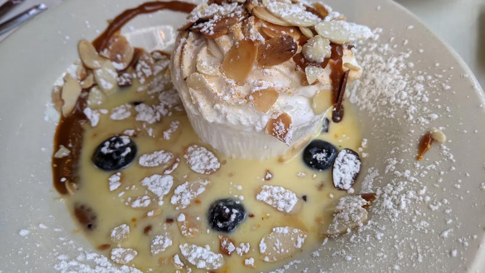 Dessert plate with cream topped with almond slices, blueberries, powdered sugar, and custard sauce drizzle.