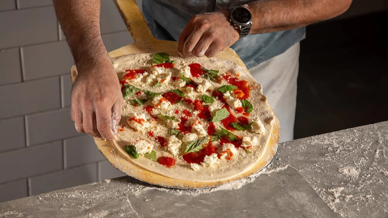 Person preparing homemade pizza with tomato sauce, cheese, and basil on a wooden paddle.