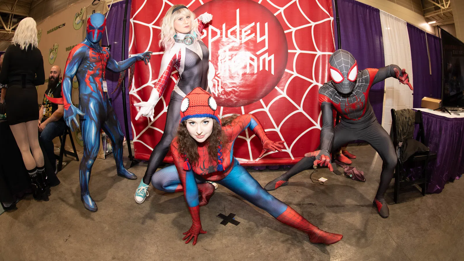 Four cosplayers dressed as Spider-Man characters pose dynamically in front of a red spider web backdrop at a convention.