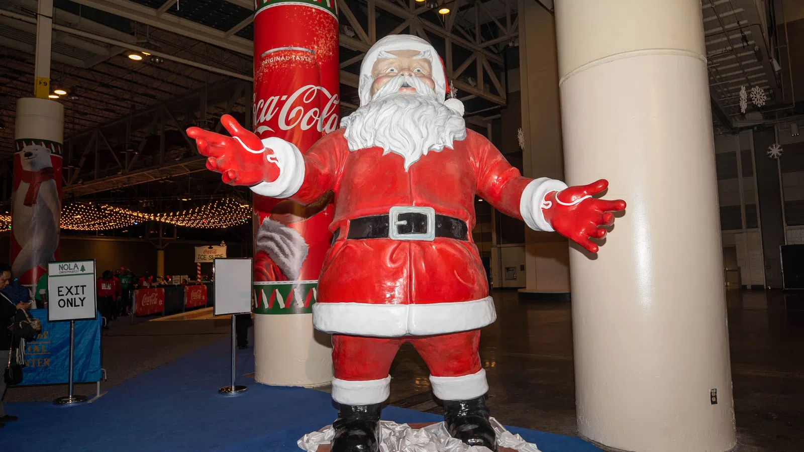 Life-size Santa Claus statue with open arms near Coca-Cola branded pillar at an indoor event.