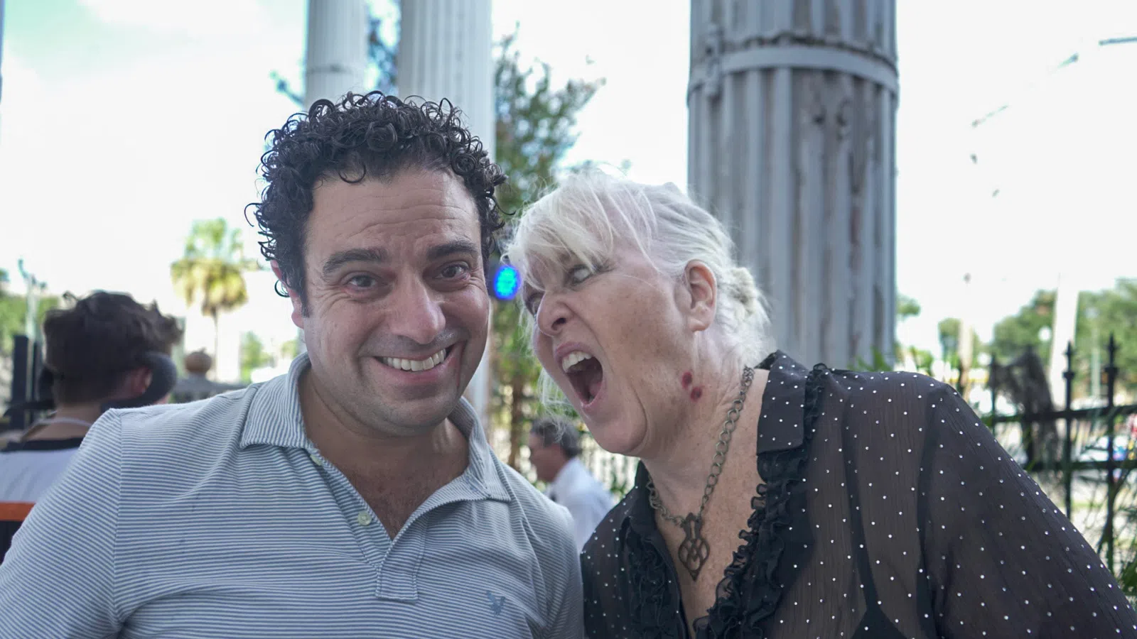 Smiling man and woman with open mouth caught mid-shout outdoors by stone columns and greenery in background