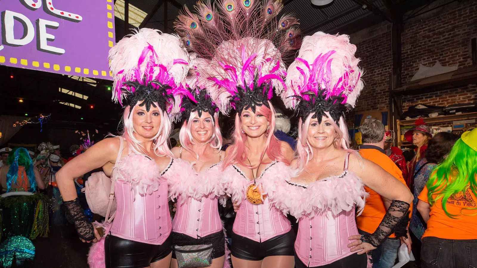 Four women in pink feathered showgirl costumes with black accents posing indoors at a festive event.