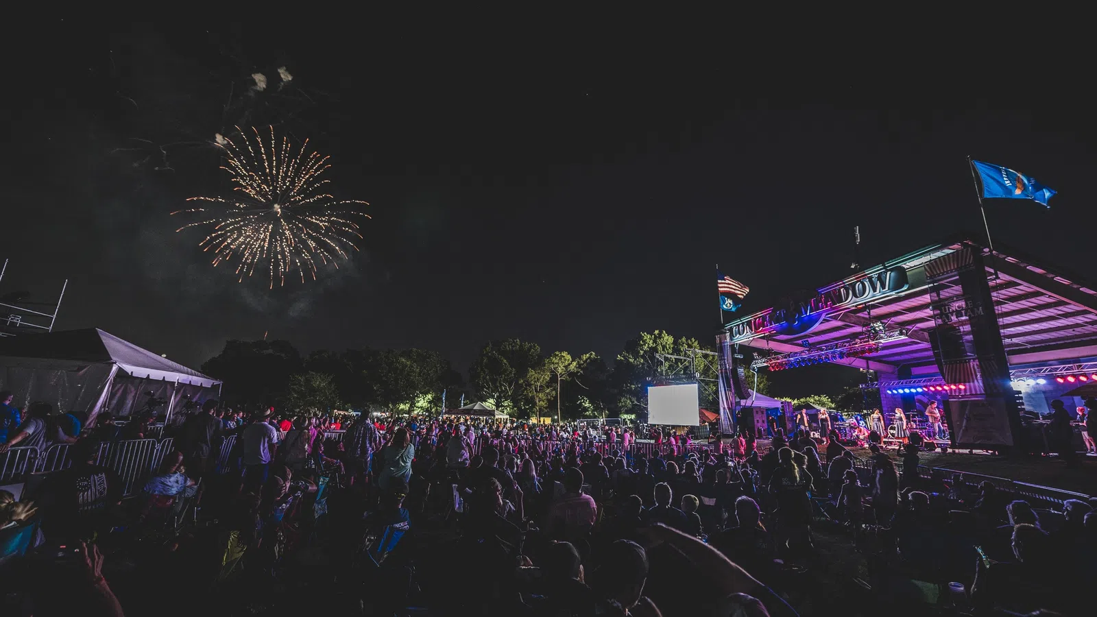 Crowd at outdoor night concert with fireworks exploding in the sky above the stage and audience