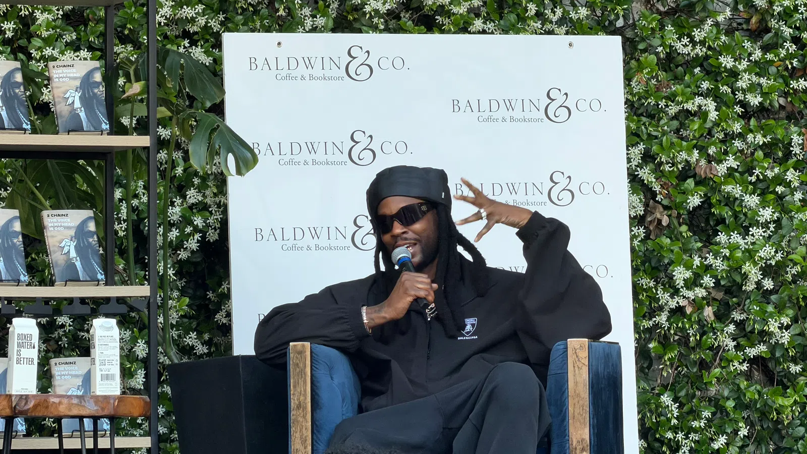 Man in black clothing and sunglasses speaking into microphone at Baldwin & Co. Coffee & Bookstore event.
