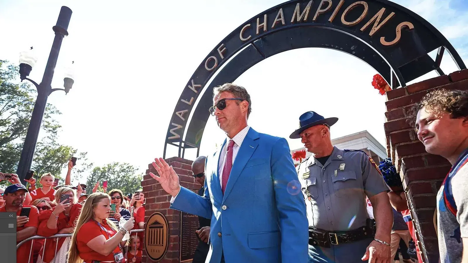 Man in blue suit walking under Walk of Champions arch with crowd and police officer nearby
