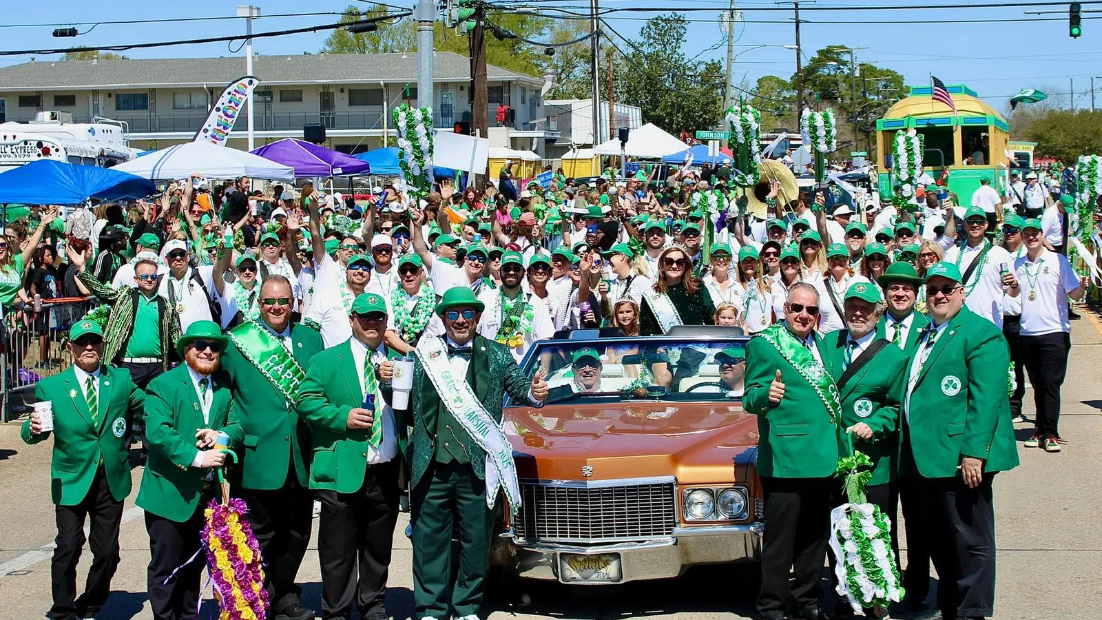 Group dressed in green suits and hats posing in front of a classic car during a festive St. Patrick's Day parade.