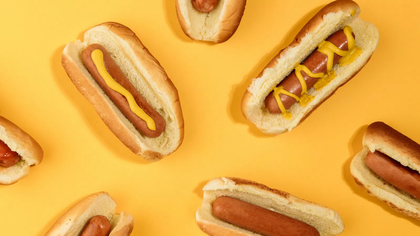Several hot dogs in buns on a yellow background, some topped with yellow mustard in various patterns.