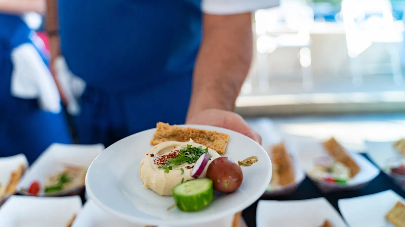 Chef holding plate with hummus, pita bread, cucumber slice, grape tomato, and garnish in a kitchen setting