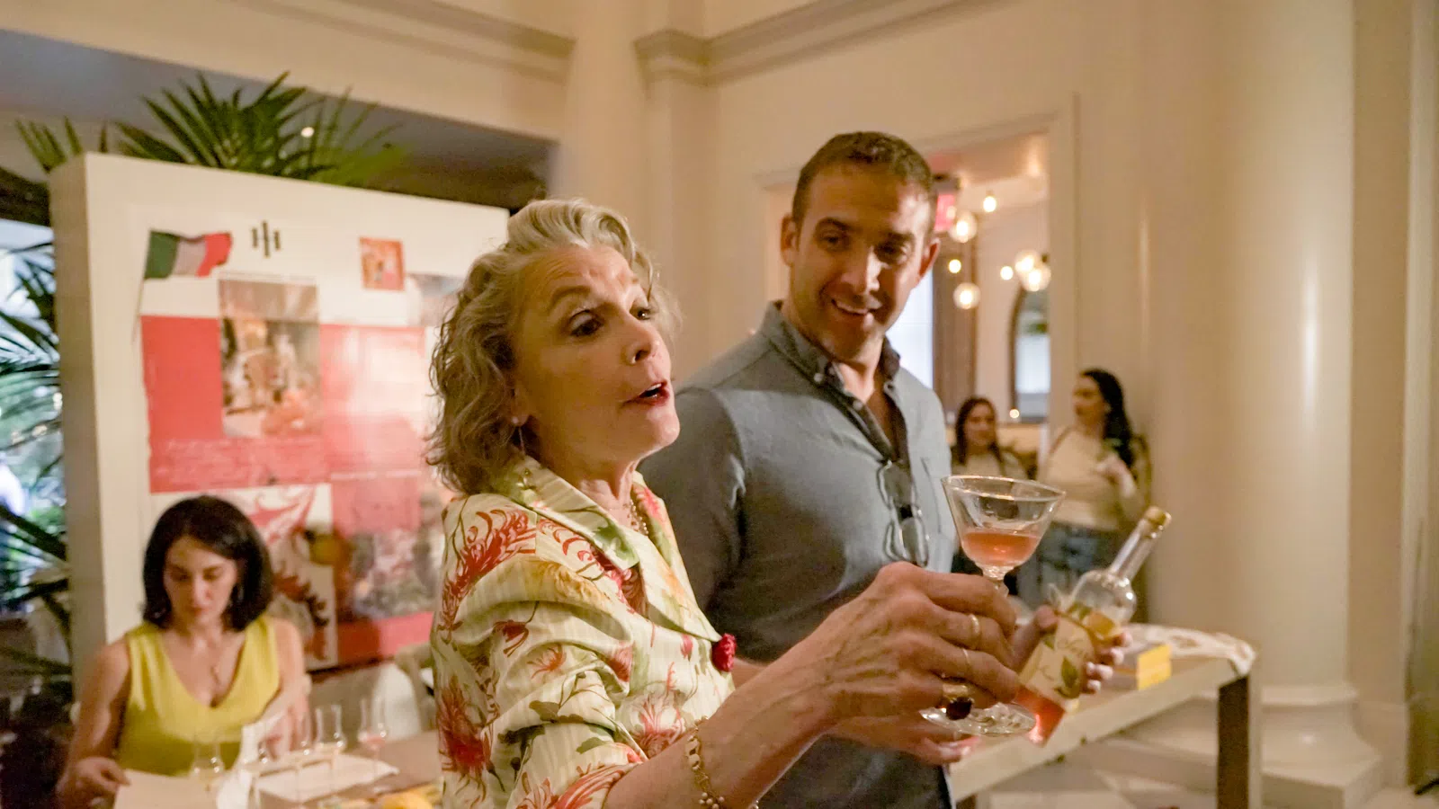Older woman pouring wine while smiling man watches in elegant room with guests and decorations