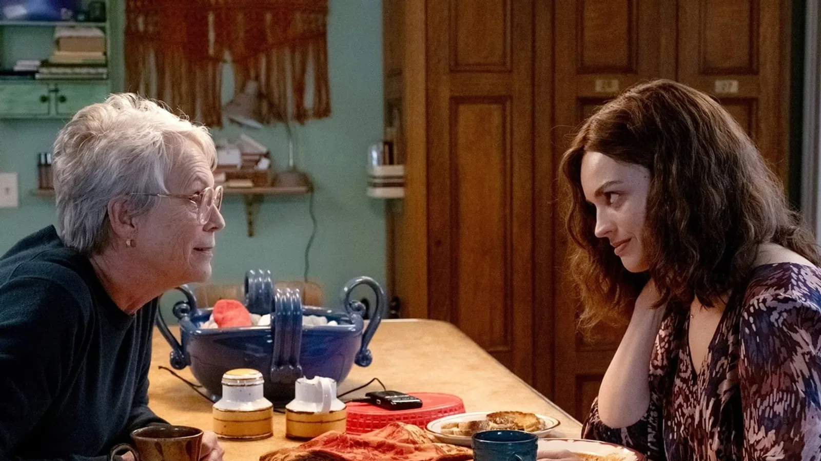 Two women sharing a warm conversation over coffee at a cozy kitchen table with plates and a large bowl.